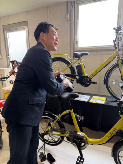 電動アシスト自転車「Rafoot」の特長について説明する佐々木社長。手に持つバッテリーはスマートフォン等の充電にも利用できる。